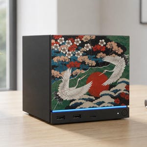 May include: A black rectangular electronic device featuring a front panel with a graphic print of two white cranes, a large red sun, and flowering branches. The base includes two USB ports, a small indicator light, and a circular power button.