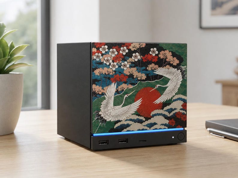 May include: A black rectangular electronic device featuring a front panel with a graphic print of two white cranes, a large red sun, and flowering branches. The base includes two USB ports, a small indicator light, and a circular power button.