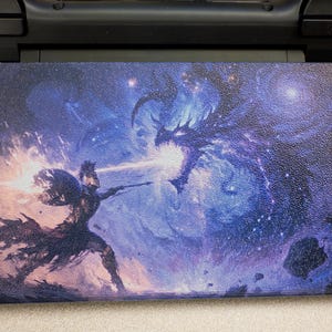 May include: A rectangular gaming console skin featuring a vibrant, detailed illustration of a warrior in armor wielding a glowing weapon against a large, dark, ethereal dragon. The scene is set against a deep blue, cosmic, starry nebula background.