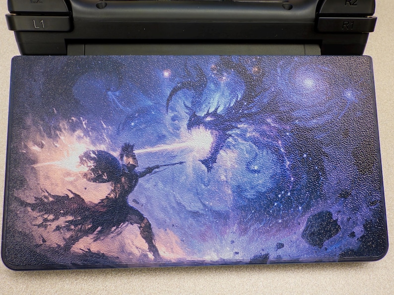 May include: A rectangular gaming console skin featuring a vibrant, detailed illustration of a warrior in armor wielding a glowing weapon against a large, dark, ethereal dragon. The scene is set against a deep blue, cosmic, starry nebula background.
