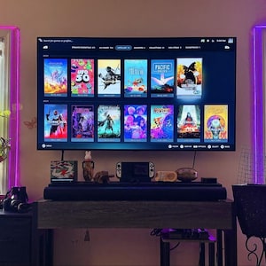 May include: A large flat-screen television mounted on a wall displays a digital game library interface with multiple colorful game cover art tiles. Below the screen sits a dark horizontal soundbar on a wooden console table with a handheld gaming device.