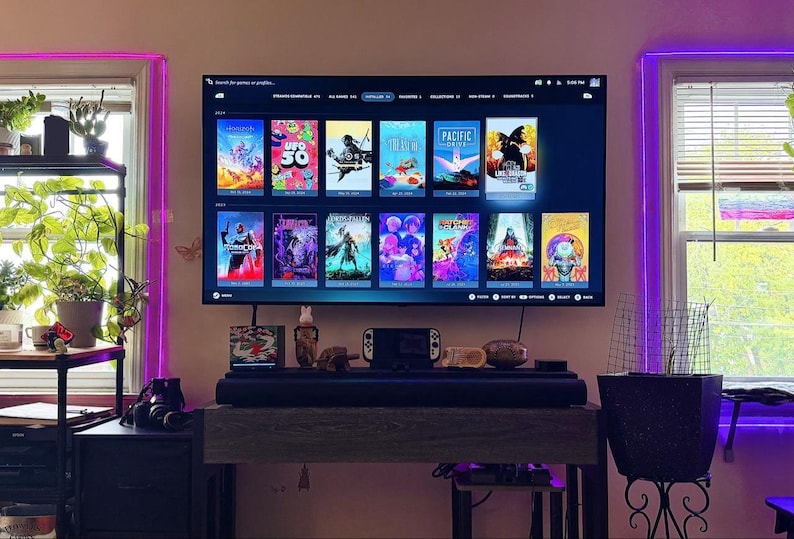 May include: A large flat-screen television mounted on a wall displays a digital game library interface with multiple colorful game cover art tiles. Below the screen sits a dark horizontal soundbar on a wooden console table with a handheld gaming device.