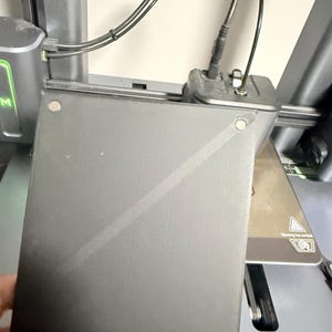 May include: A square black 3D printer build plate with a textured surface, featuring small circular magnetic attachment points at the corners and a diagonal line across the center, positioned in front of a dark gray 3D printer assembly.