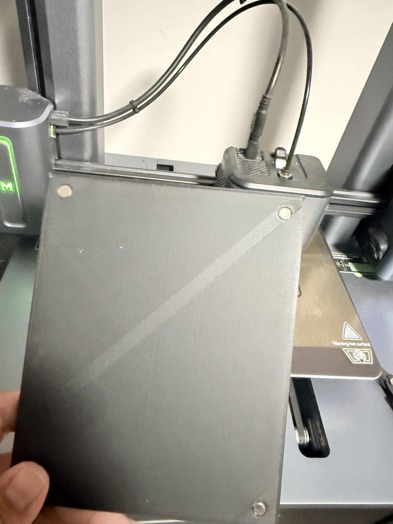 May include: A square black 3D printer build plate with a textured surface, featuring small circular magnetic attachment points at the corners and a diagonal line across the center, positioned in front of a dark gray 3D printer assembly.