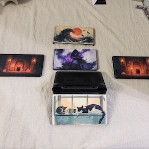May include: A black handheld gaming console with a rectangular screen cover featuring a cat illustration, surrounded by four additional rectangular screen covers displaying various artistic designs including a wave, a hooded figure, and dungeon scenes.