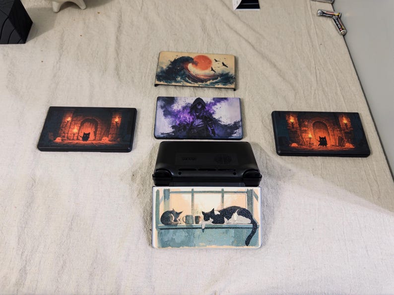 May include: A black handheld gaming console with a rectangular screen cover featuring a cat illustration, surrounded by four additional rectangular screen covers displaying various artistic designs including a wave, a hooded figure, and dungeon scenes.