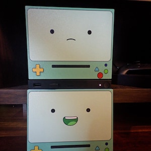May include: A black electronic console featuring two light green panels decorated with the BMO character from Adventure Time. The top panel shows a neutral face, while the bottom panel displays a smiling face with a green mouth and white teeth.