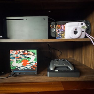 May include: A black gaming console, a handheld gaming device, and a white instant camera with a wrist strap on the top shelf. A black box with a colorful crane and cherry blossom graphic and a gray game controller on a charging dock below.