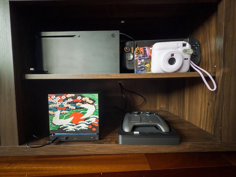 May include: A black gaming console, a handheld gaming device, and a white instant camera with a wrist strap on the top shelf. A black box with a colorful crane and cherry blossom graphic and a gray game controller on a charging dock below.