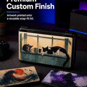 May include: A black handheld gaming console with a snap-fit lid featuring a printed illustration of two cats resting on a windowsill. Two additional rectangular lids are displayed in the foreground, one with a wave and sun design and one with a purple abstract scene.