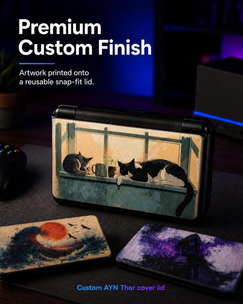 May include: A black handheld gaming console with a snap-fit lid featuring a printed illustration of two cats resting on a windowsill. Two additional rectangular lids are displayed in the foreground, one with a wave and sun design and one with a purple abstract scene.