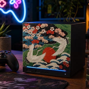 May include: A black square gaming console featuring a decorative front panel with a detailed illustration of two white cranes, a large red sun, stylized blue waves, and pink cherry blossoms. The device has a thin horizontal light strip at the base.