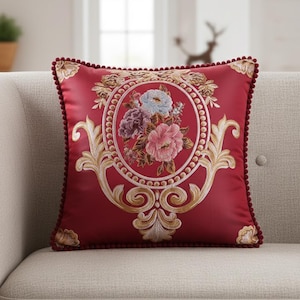Peut inclure: Un coussin carré bordeaux avec un motif floral brodé. Le centre présente un bouquet de fleurs roses, bleues et violettes dans un cadre circulaire. Des volutes dorées et une bordure en pompons rouge foncé complètent le coussin décoratif.