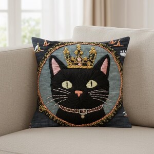 Peut inclure: Coussin carré brodé avec un chat noir portant une couronne et un collier dorés. Le chat est centré sur un fond bleu sarcelle, encadré par une bordure décorative. Le coussin est sur un canapé beige.