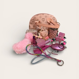 Può includere: Una collezione di accessori rosa, tra cui una piccola borsa, un rossetto, un portachiavi e un allarme di sicurezza personale. Gli articoli sono disposti su una superficie bianca, con uno sfondo morbido e chiaro.