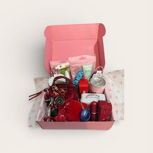 Könnte beinhalten: Eine rosa Geschenkbox gefüllt mit verschiedenen kleinen Gegenständen. Die Box enthält eine rote Handtasche, Schlüsselanhänger, eine kleine Dose, einen Lippenstift und anderes Zubehör. Die Artikel sind auf Seidenpapier mit rosa Schleifen angeordnet.