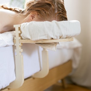 Può includere: Un poggiatesta per lettino da massaggio in legno con un cuscino bianco e una persona sdraiata a faccia in giù. Il poggiatesta è realizzato in legno chiaro ed è progettato per essere fissato a un telaio del letto. Il cuscino è in tessuto bianco e morbido.