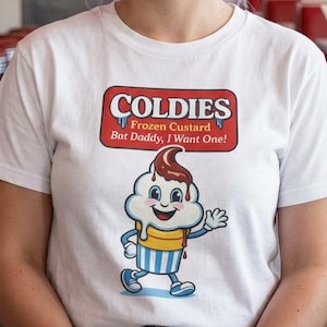 May include: White t-shirt with a "Coldies" logo featuring a cartoon ice cream cone. The logo is in a red and white rectangular design with the text "Frozen Custard But Daddy, I Want One!" The person is smiling in a diner setting.