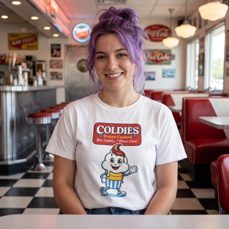 May include: White t-shirt with a "Coldies" logo featuring a cartoon ice cream cone. The logo is in a red and white rectangular design with the text "Frozen Custard But Daddy, I Want One!" The person is smiling in a diner setting.
