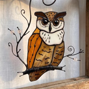 May include: A stained glass owl suncatcher featuring amber and brown textured glass wings, a white chest with black detailing, and dark glass eyes. The owl is perched on a black metal branch frame with decorative swirls and small pink beads.