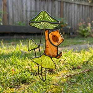 Puede incluir: Una estaca de jard&iacute;n de vidrieras con un gato atigrado naranja colgando de una seta verde. El gato tiene ojos marrones y est&aacute; sobre un fondo de hierba verde y una valla de madera. La seta tiene un sombrero verde y un tallo blanco.