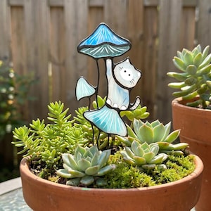 Puede incluir: Una estaca de jard&iacute;n caprichosa con un gato blanco agarrado a una seta azul. El gato y la seta son de vidrieras. La estaca est&aacute; colocada en una maceta de terracota llena de suculentas y musgo.