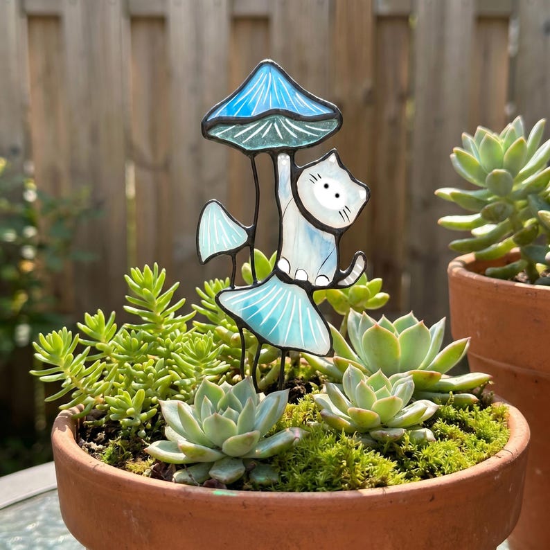 Puede incluir: Una estaca de jard&iacute;n caprichosa con un gato blanco agarrado a una seta azul. El gato y la seta son de vidrieras. La estaca est&aacute; colocada en una maceta de terracota llena de suculentas y musgo.
