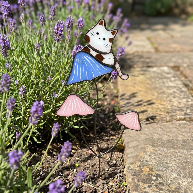 Puede incluir: Una estaca de jard&iacute;n caprichosa con un gato blanco con manchas marrones posado sobre un elemento en forma de hongo azul. Debajo, dos acentos de hongos rosas a&ntilde;aden un toque juguet&oacute;n. La estaca est&aacute; colocada entre plantas de lavanda.