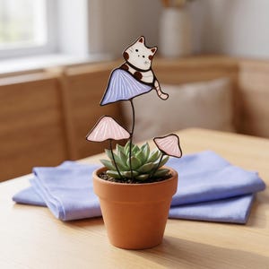 Puede incluir: Una maceta de terracota contiene una planta suculenta, adornada con una estaca de jard&iacute;n de gato y hongo. El gato es blanco con manchas marrones, posado sobre un hongo azul. Los hongos rosas acent&uacute;an la decoraci&oacute;n.