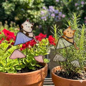 Puede incluir: Dos estacas de jard&iacute;n de vidrio manchado con gatos y acentos de hongos. Un gato es blanco con manchas marrones y una falda azul, el otro es naranja con manchas marrones y una falda verde. Las estacas est&aacute;n en macetas de terracota con flores rojas.