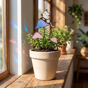 Puede incluir: Una maceta de terracota con una planta y estacas decorativas para jard&iacute;n. Las estacas representan un gato blanco con manchas marrones sobre una seta azul y dos setas rosas. La maceta est&aacute; sobre un alf&eacute;izar de madera.