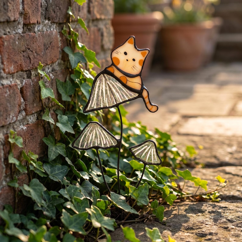 Puede incluir: Una estaca de jard&iacute;n caprichosa con un gato posado sobre una seta. El gato tiene detalles en naranja y blanco, y las setas tienen un dise&ntilde;o a rayas. La estaca est&aacute; rodeada de hiedra verde y colocada contra una pared de ladrillo.