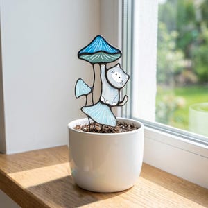 Puede incluir: Una estaca decorativa para plantas de vidrieras que representa un gato gris claro sobre una seta azul. La seta tiene un sombrero azul y est&aacute; sostenida por un tallo y dos sombreros azules m&aacute;s peque&ntilde;os. La estaca est&aacute; en una maceta de cer&aacute;mica blanca.