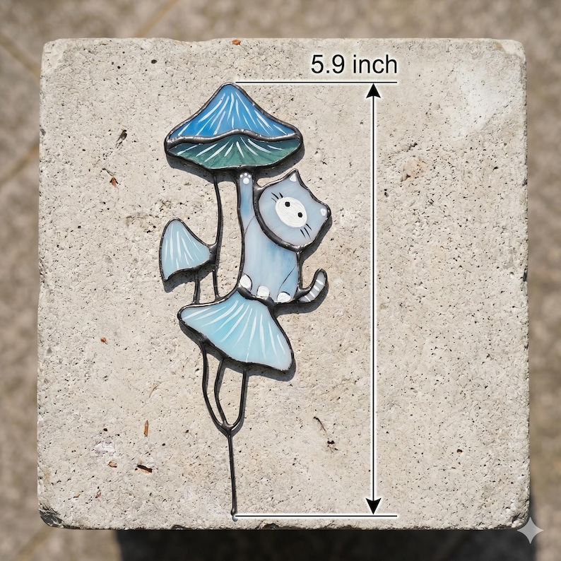Puede incluir: Una estaca de jard&iacute;n caprichosa con un gato azul claro agarrado a una seta con una tapa azul y verde. La estaca mide 15 cm de alto, con un dise&ntilde;o decorativo de seta y gato.