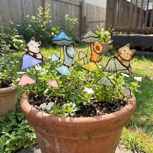 Puede incluir: Una maceta de terracota llena de vegetaci&oacute;n y flores blancas, con estacas de jard&iacute;n caprichosas de gatos y setas. Las estacas tienen coloridas tapas de setas de cristal y adorables figuras de gatos en varias poses, a&ntilde;adiendo un toque l&uacute;dico a la decoraci&oacute;n exterior.