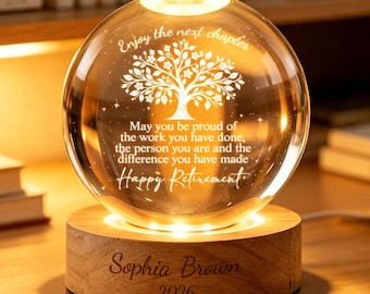 Puede incluir: Bola de cristal con diseño de árbol y las palabras "Enjoy the next chapter" y "Happy Retirement" grabadas. Se apoya en una base de madera con el nombre "Sophia Brown" y el año "2026" grabados. La base está iluminada.
