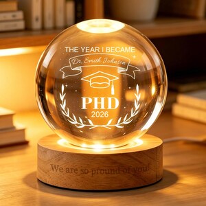 May include: A clear glass sphere engraved with "THE YEAR I BECAME" and "Dr. Smith Johnson", a graduation cap, and "PHD 2026". The sphere sits on a wooden base with the text "We are so proud of you!" and is illuminated with warm light.