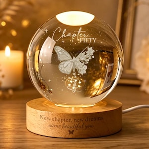Op de afbeelding: Een nachtlampje van een heldere glazen bol op een ronde houten voet. De bol is voorzien van een gegraveerde vlinder en de tekst "Chapter FIFTY". De houten voet is gegraveerd met de tekst "New chapter, new dreams same beautiful you" en een kleine vlinder.