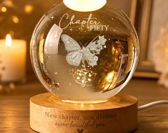 Puede incluir: Una luz nocturna de esfera de cristal transparente sobre una base de madera redonda. La esfera presenta una mariposa grabada y el texto "Chapter FIFTY". La base de madera está grabada con el texto "New chapter, new dreams same beautiful you" y una pequeña mariposa.