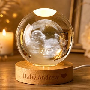 Può includere: Una sfera di vetro trasparente con un'immagine ecografica all'interno, appoggiata su una base di legno. La base è incisa con la scritta "Baby Andrew" e un cuore. La sfera è illuminata dall'interno, creando un bagliore caldo.