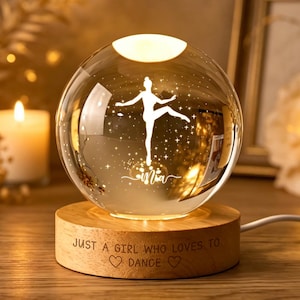 Puede incluir: Una luz nocturna de esfera de cristal transparente con una silueta blanca grabada de una bailarina y el nombre "Mia" en su interior. La esfera descansa sobre una base redonda de madera grabada con el texto "JUST A GIRL WHO LOVES TO DANCE" y dos corazones.
