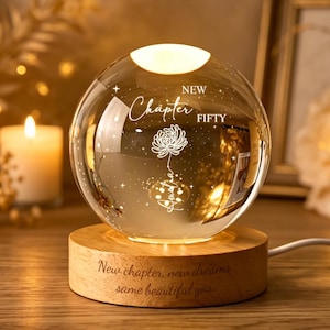 Op de afbeelding: Een heldere glazen bolvormige lamp op een ronde houten voet. De bol is voorzien van een gegraveerd bloemmotief met de tekst "NEW CHAPTER FIFTY". De houten voet is gegraveerd met de tekst "New chapter, new dreams, same beautiful you."