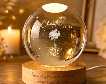 Puede incluir: Una lámpara de esfera de cristal transparente sobre una base redonda de madera. La esfera presenta un diseño floral grabado con el texto "NEW CHAPTER FIFTY". La base de madera tiene grabado el texto "New chapter, new dreams, same beautiful you."