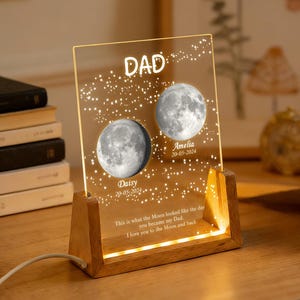 Op de afbeelding: Een rechthoekige, doorzichtige acrylplaat in een lichte houten voet met geïntegreerde ledverlichting. De plaat bevat de tekst DAD, twee gedetailleerde maanillustraties met de namen Daisy en Amelia, datums en de zin I love you to the moon and back.
