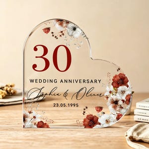May include: A heart-shaped clear acrylic plaque featuring the number 30 in dark red, the text WEDDING ANNIVERSARY, the names Sophia & Oliver in black script, and the date 23.05.1995, decorated with printed red and white floral illustrations.