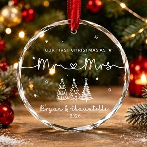 May include: A circular clear glass Christmas ornament with beveled edges, featuring a red ribbon hanger. The text reads "OUR FIRST CHRISTMAS AS Mr & Mrs", "Bryan & Chauntelle", and "2026", accompanied by three white line-art Christmas trees.