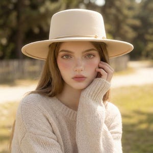 May include: A beige felt fedora hat with a flat crown and a wide brim, featuring a thin metallic gold band encircling the base of the crown. The hat is worn with a matching beige knit sweater that has a textured, chunky weave and long sleeves.