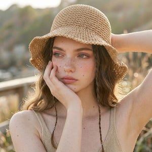 May include: A beige, crocheted sun hat with a slightly ruffled brim. The hat is made of a woven material and has a rounded crown. The hat is worn with a beige vest top and braided hair.