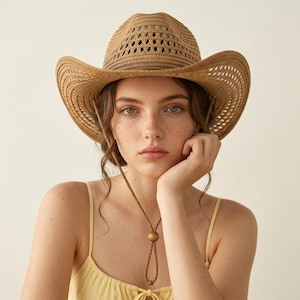 Puede incluir: Un sombrero de vaquero marrón claro con una corona perforada y un ala ancha. El sombrero tiene una correa para la barbilla con una cuenta de madera. El sombrero está hecho de material tejido. La persona lleva una camiseta sin mangas amarilla.