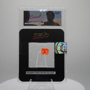 Op de afbeelding: Een beoordeelde verzamelkaart met een scène uit "Better Call Saul". De kaart zit in een transparante plastic houder met een zwarte achterkant. De houder heeft een holografische sticker en de tekst "FRAGMENT FROM 'BETTER CALL SAUL'".